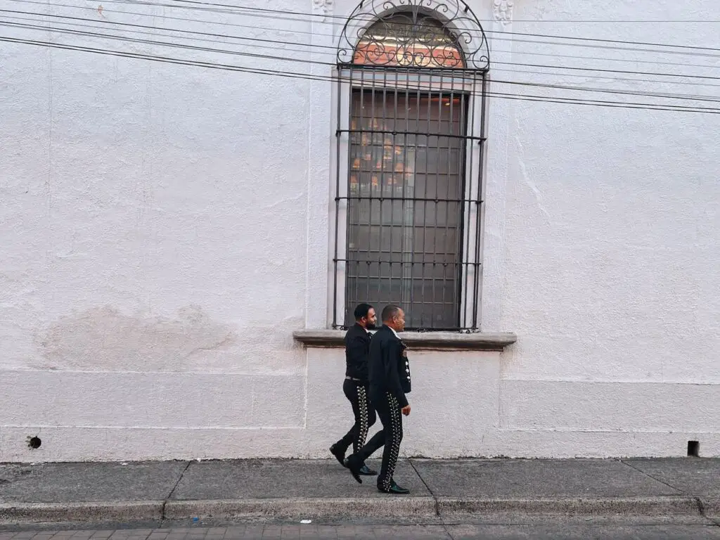 Two mariachi musicians in traditional black outfits walking down a quiet Guadalajara street.