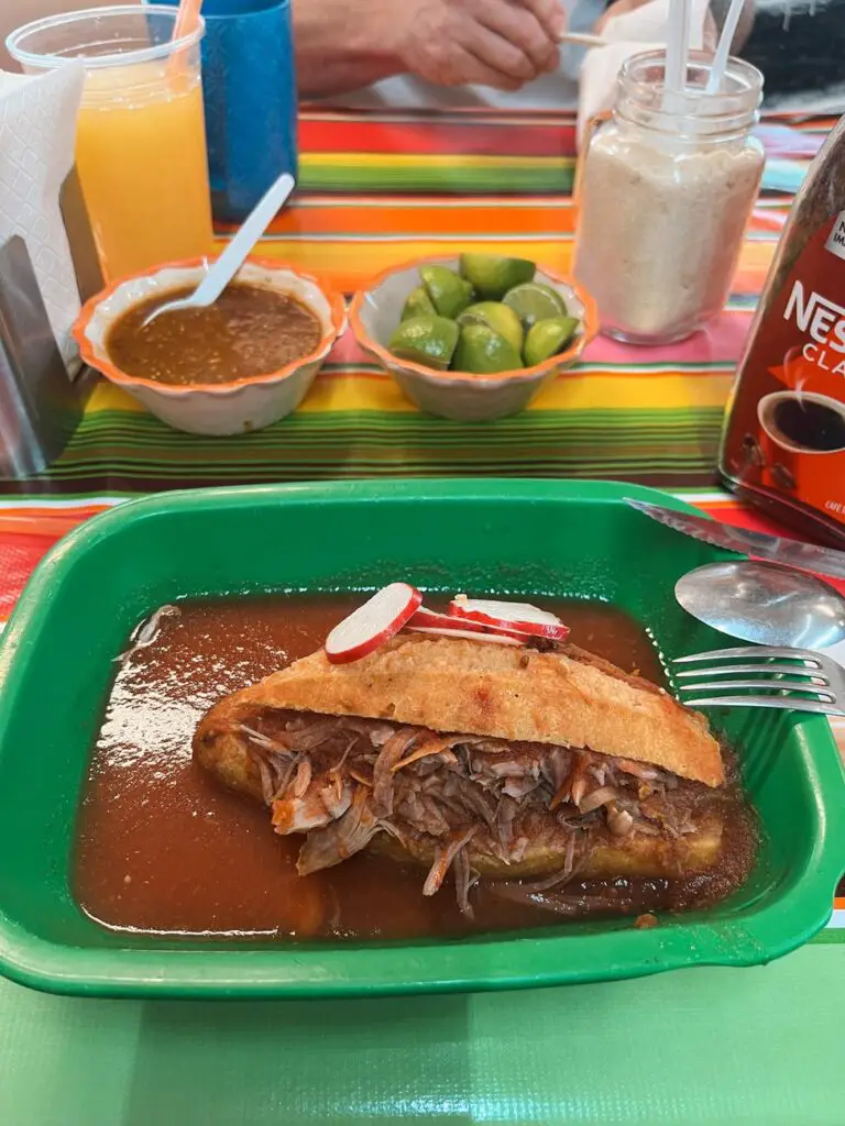 A classic torta ahogada (drowned sandwich) filled with shredded pork and smothered in spicy red sauce, served at a casual eatery with salsa, lime, and drinks on the side.