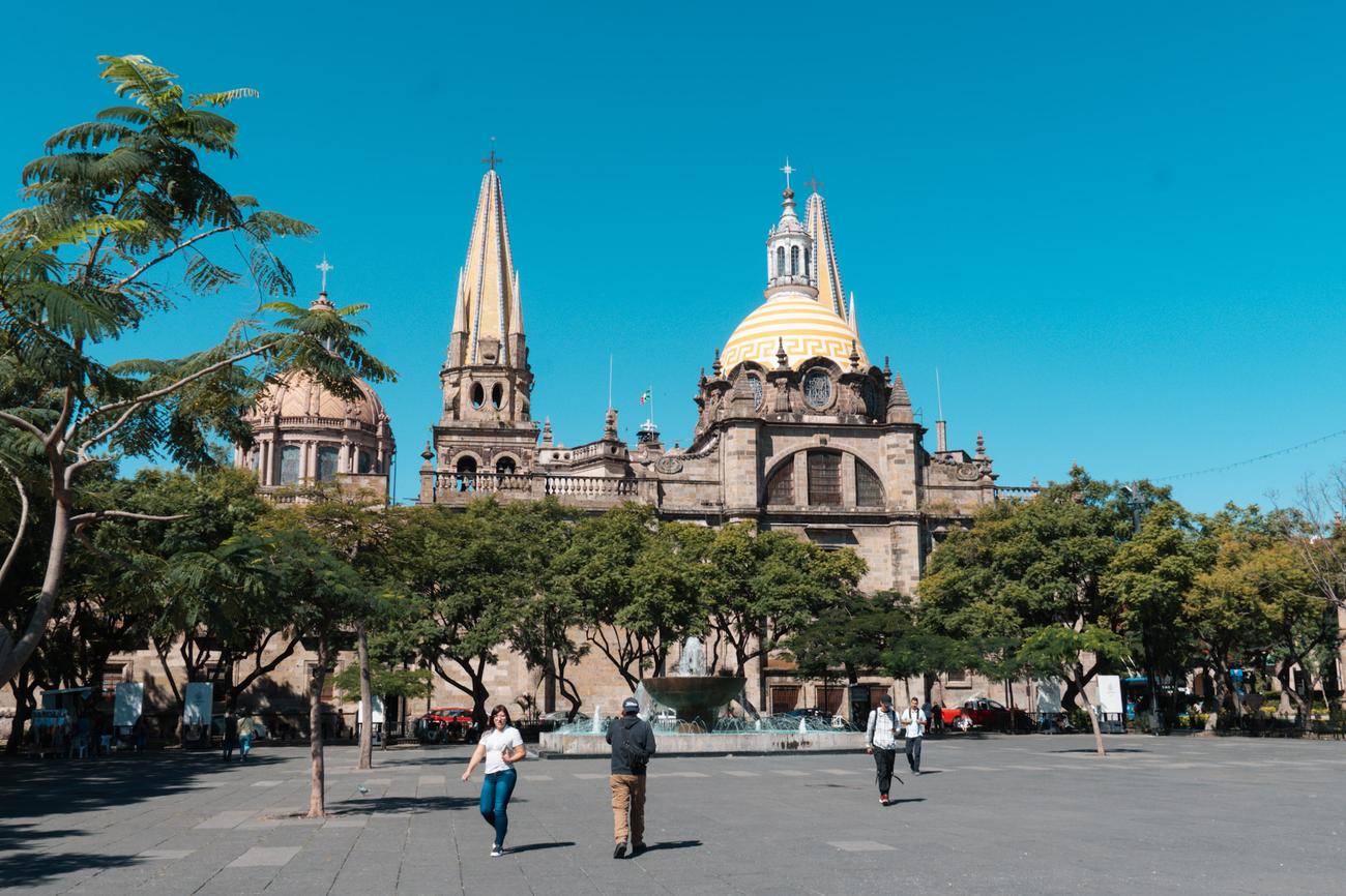 Wide view of Guadalajara Cathedral and its iconic yellow dome, seen from the main plaza (zócalo), with people walking across the square on a sunny day.