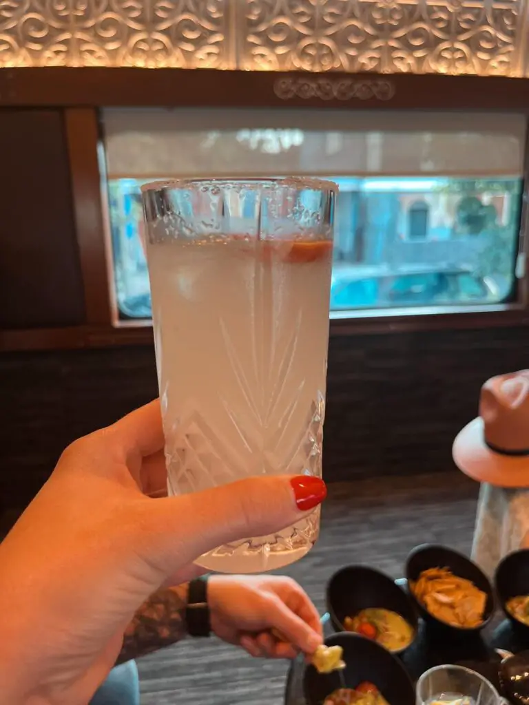 A refreshing tequila-based cocktail held up inside the Jose Cuervo Express train, with snacks and elegant decor in the background.