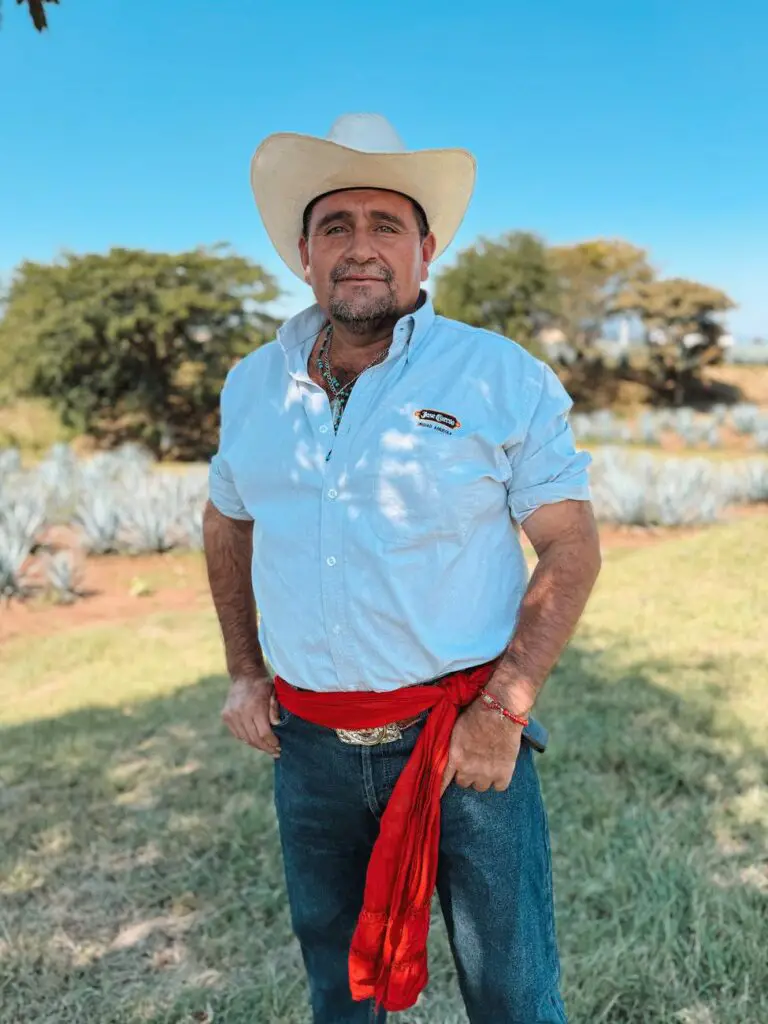 Portrait of a jimador (agave farmer) standing proudly in an agave field, wearing a white shirt, cowboy hat, and red sash, with blue sky and trees in the background.