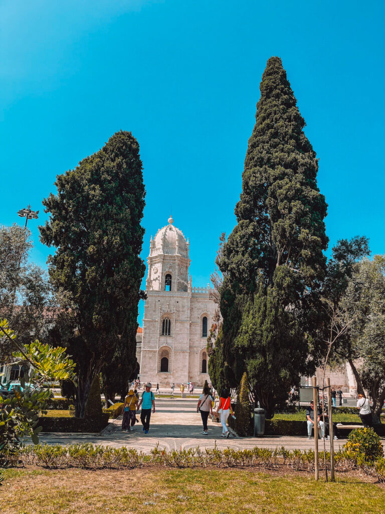 Jerónimos Monastery partially framed by tall trees in Belém, with tourists walking toward the entrance – a key historic site to visit on a 2-day Lisbon itinerary.