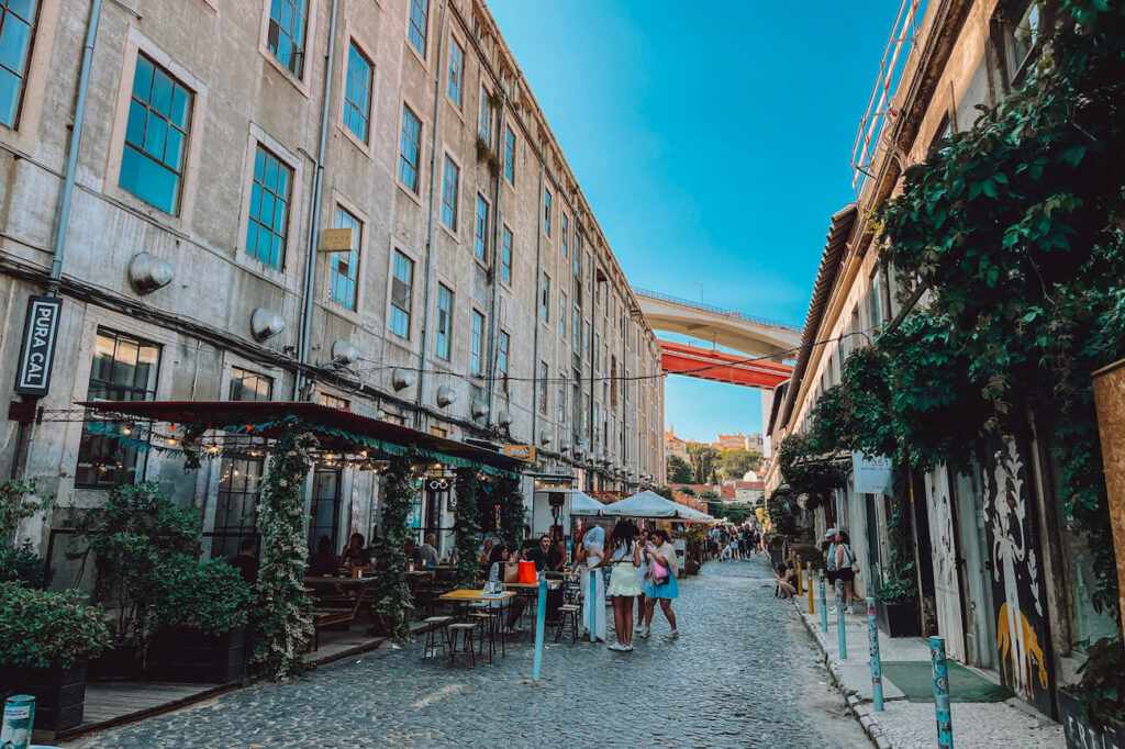 People dining at open-air restaurants along a cobblestone street in the LX Factory district, with a bridge visible above.
