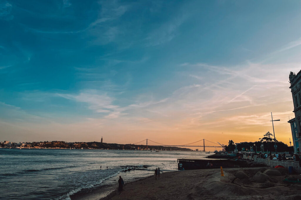 Sunset view over the Tagus River with the 25 de Abril Bridge in the distance and silhouettes of people walking along the shore – a relaxing moment to enjoy during a weekend in Lisbon.