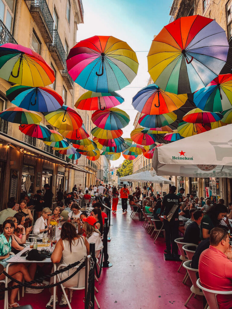 Colorful umbrellas hanging above a lively pedestrian dining street in Lisbon, with people enjoying meals below – a Lisbon's Pink Street is a popular place for nightlife.
