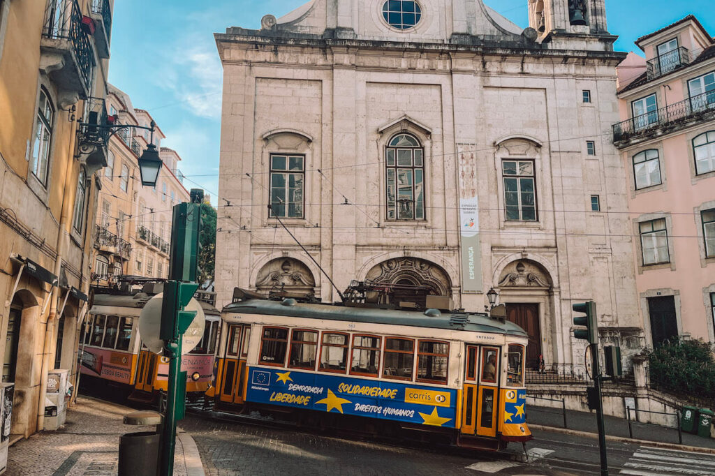 Classic yellow trams passing through a historic Lisbon neighbourhood in front of a stone church – an iconic experience to include in your 2-day Lisbon itinerary.