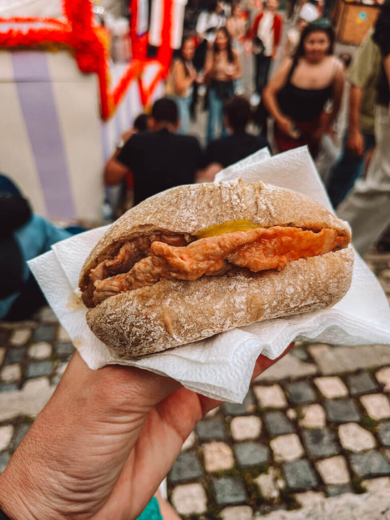 Close-up of a bifana, a traditional Portuguese pork sandwich held in hand during a local event – a delicious street food to try during a weekend in Lisbon.