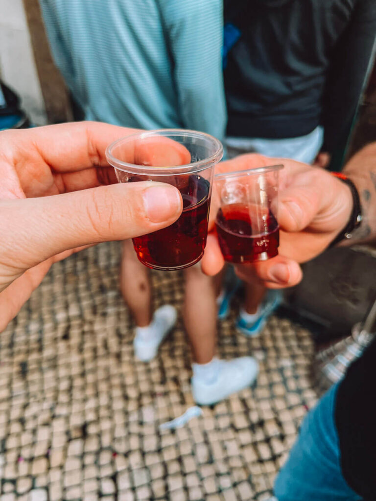 Two hands holding small plastic cups of Ginjinha, a traditional cherry liqueur – a must-try local drink when visiting Lisbon.