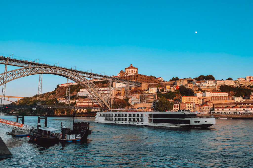 Panoramic view of the Dom Luís I Bridge and Douro River with boats and a large river cruise ship at sunset – a classic sight to enjoy during a weekend in Porto.