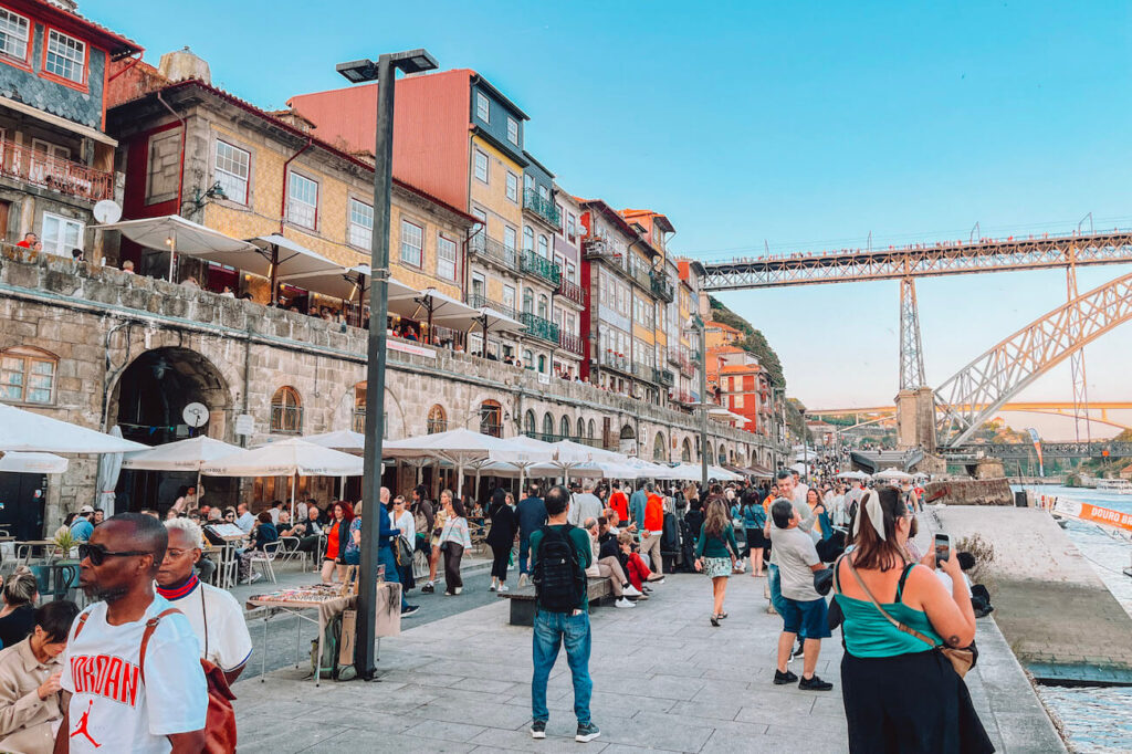 Crowds enjoying riverside restaurants and cafes along Cais da Ribeira, with colourful buildings and the Dom Luís I Bridge in the background – a lively waterfront to visit in Porto.