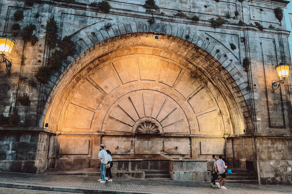 Illuminated stone arch of Fonte das Virtudes in Porto, with people passing by in the evening – a quiet, atmospheric spot worth a stop on a Porto itinerary.