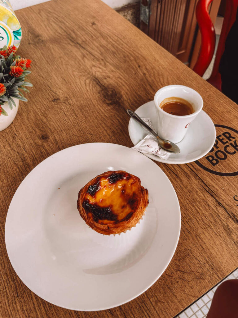 A pastel de nata (Portuguese custard tart) on a white plate beside a small cup of espresso on a wooden café table, capturing a typical pilgrim’s snack break.