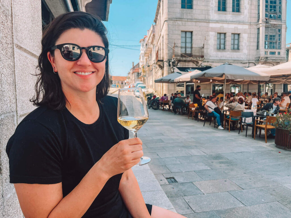 A woman - the author of this article - smiling and holding a glass of white wine while sitting outside a stone building, with a bustling street café in the background. She's in Tui, Spain.