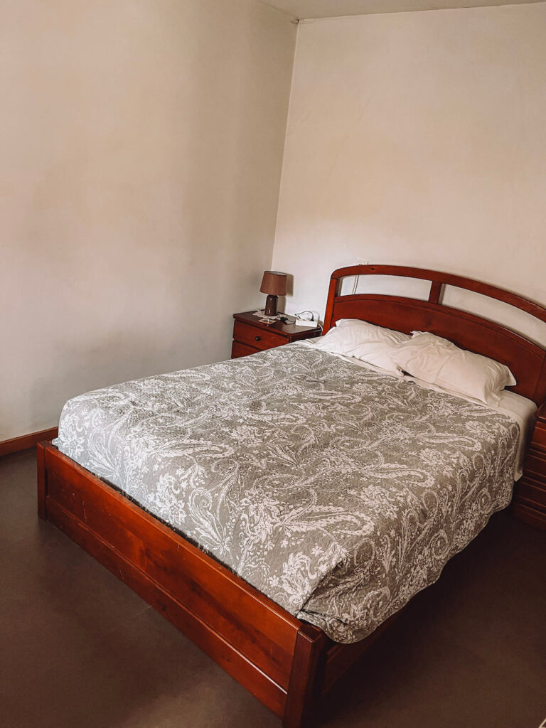 A simple bedroom with a double bed covered in a patterned duvet, flanked by wooden nightstands and a lamp - a typical, simple pilgrim’s accommodation.