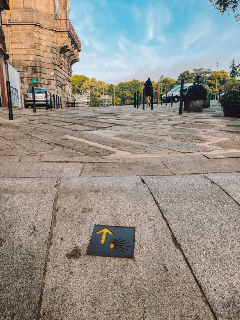 A small tile embedded in a cobblestone street with a yellow arrow and scallop shell symbol, indicating the Portuguese Camino route through an urban setting.