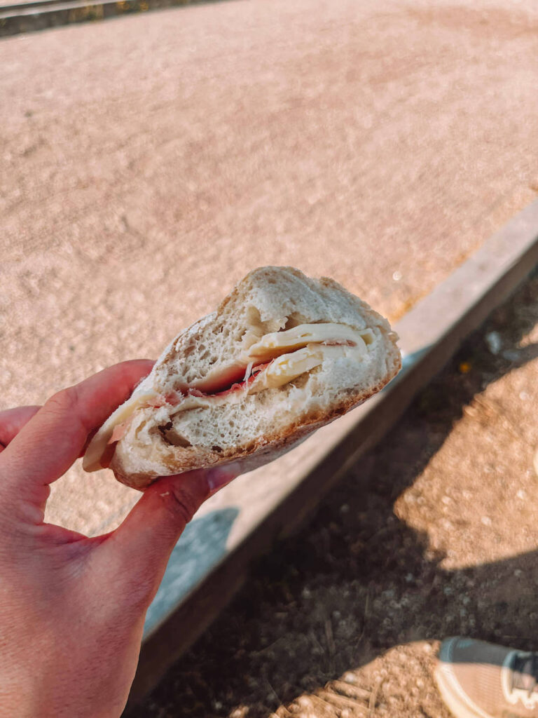 A close-up of a hand holding a simple sandwich with ham and cheese on crusty bread, with a gravel ground and foot partially visible in the background.