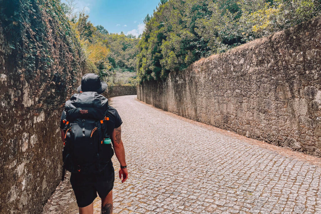 A solo hiker with a black backpack walking along a cobblestone road flanked by high stone walls and lush greenery, following the Camino path.