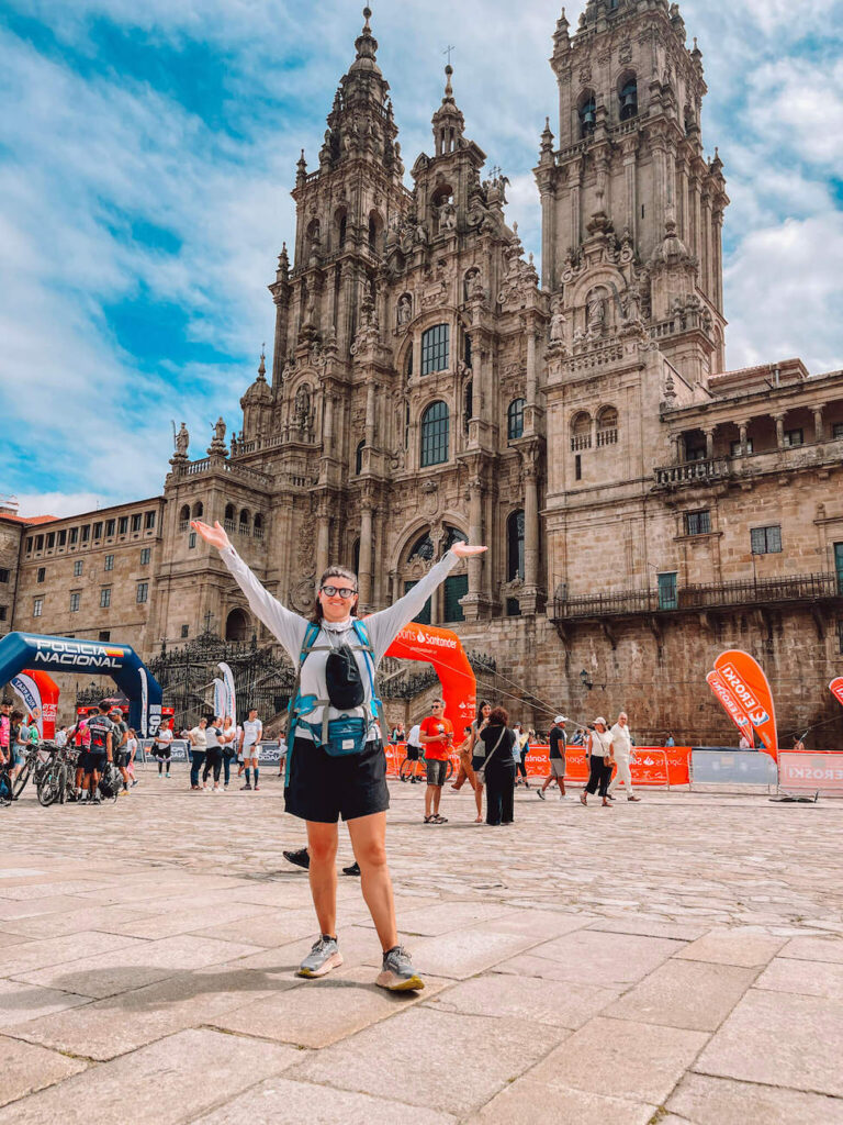 A triumphant pilgrim - the author of this article - stands with arms raised in front of the Santiago de Compostela Cathedral, surrounded by other walkers and celebratory signage.