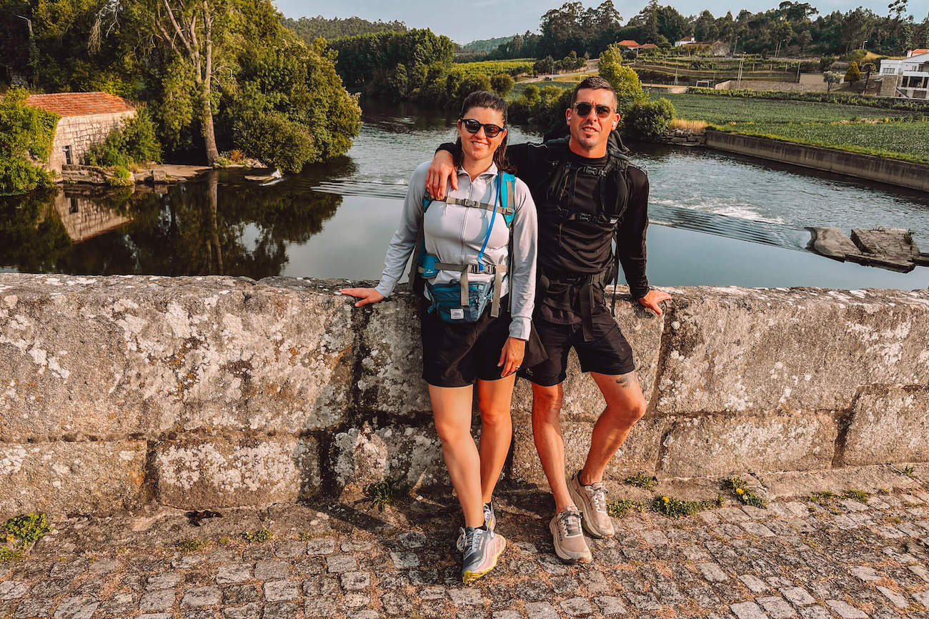 Two hikers - the author and her husband - dressed in activewear standing on a stone bridge on the Portuguese Camino, smiling at the camera with a river and green countryside in the background.