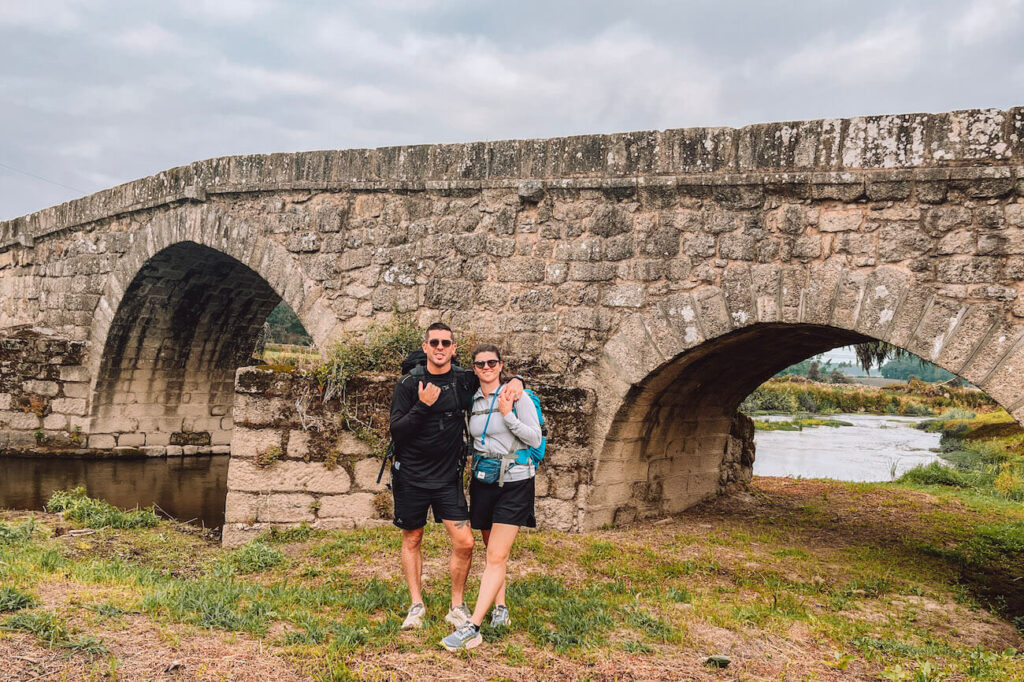 Two hikers - the author and her husband - pose in front of a stone medieval bridge, wearing backpacks and activewear suitable for the Portuguese Camino.