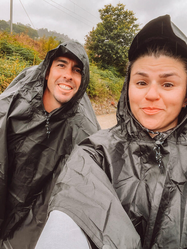 A couple - the author and her husband - wearing black rain ponchos smiles during wet weather on the trail, showing essential waterproof gear for the Portuguese Camino.