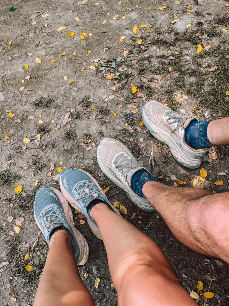Two pairs of legs and trail shoes are shown resting on a dirt path - good shoes with thick soles are essential for your Portuguese Camino packing list.