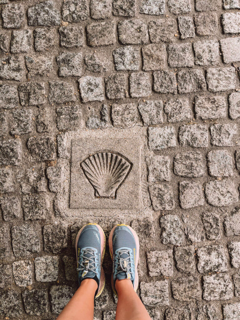 A pair of feet in trail runners stands next to a scallop shell symbol embedded in cobblestones, highlighting appropriate footwear for the Portuguese Camino.