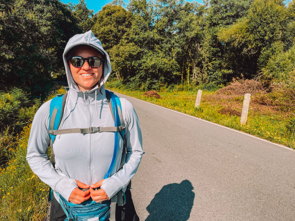 The author in sunglasses and a hooded long-sleeve shirt stands on a rural road - sun protection clothing and a good daypack are Portuguese Camino packing list essentials.