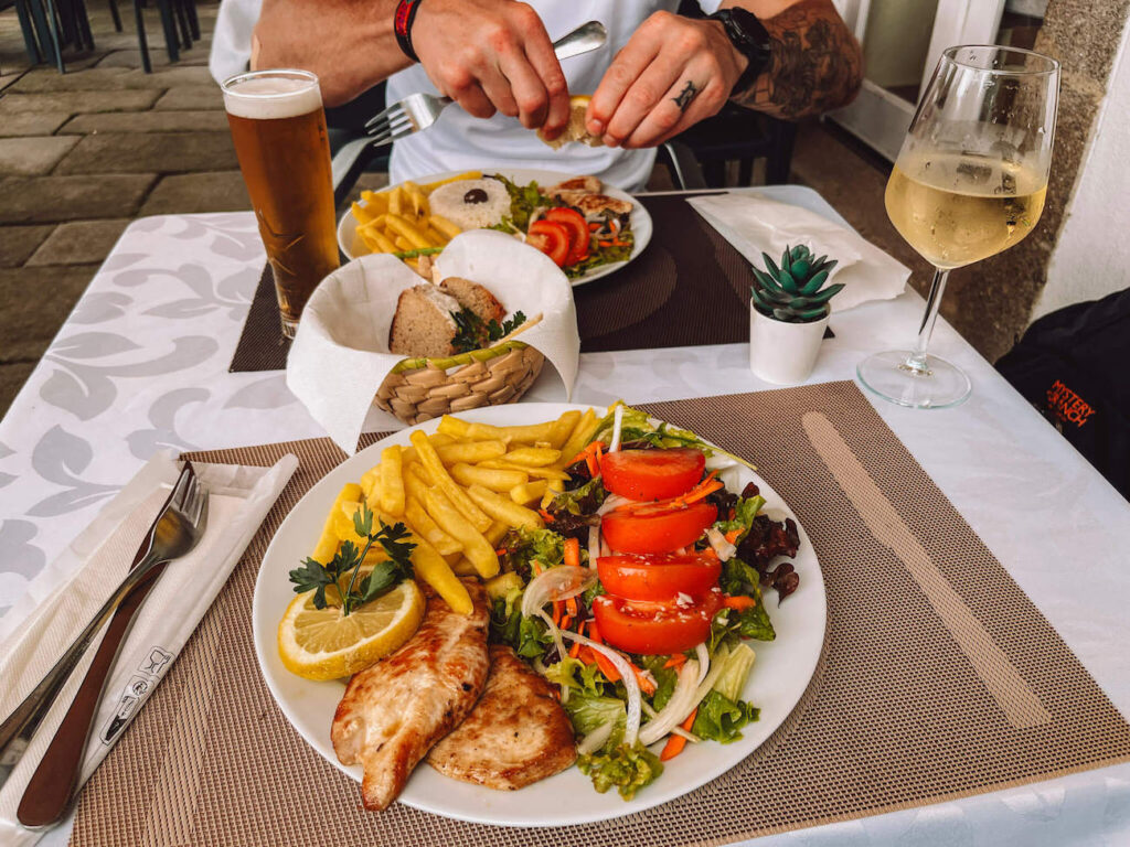 A plate of grilled chicken, French fries, and fresh salad served at an outdoor restaurant, with a glass of beer, a glass of white wine, and a bread basket on the table.