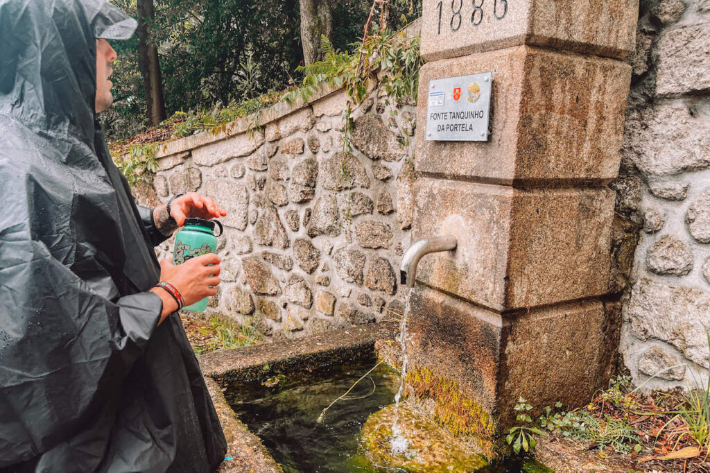 A pilgrim in a rain poncho filling a water bottle from a stone fountain on the Portuguese Camino labelled “Fonte Tanquinho da Portela,” built into a mossy stone wall.