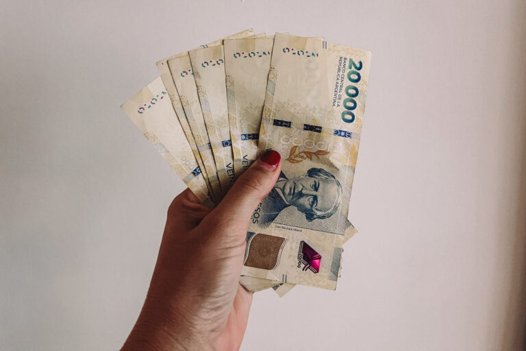 A hand - the author's - holding Argentine pesos against a white wall. Money in Argentina can be confusing for travellers!