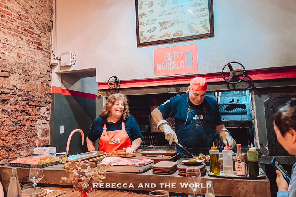 Open kitchen scene at Betty and Marcelo Buenos Aires, showing a man grilling meat and a woman smiling beside him behind a counter, capturing the warm, interactive atmosphere of a family asado in Buenos Aires.