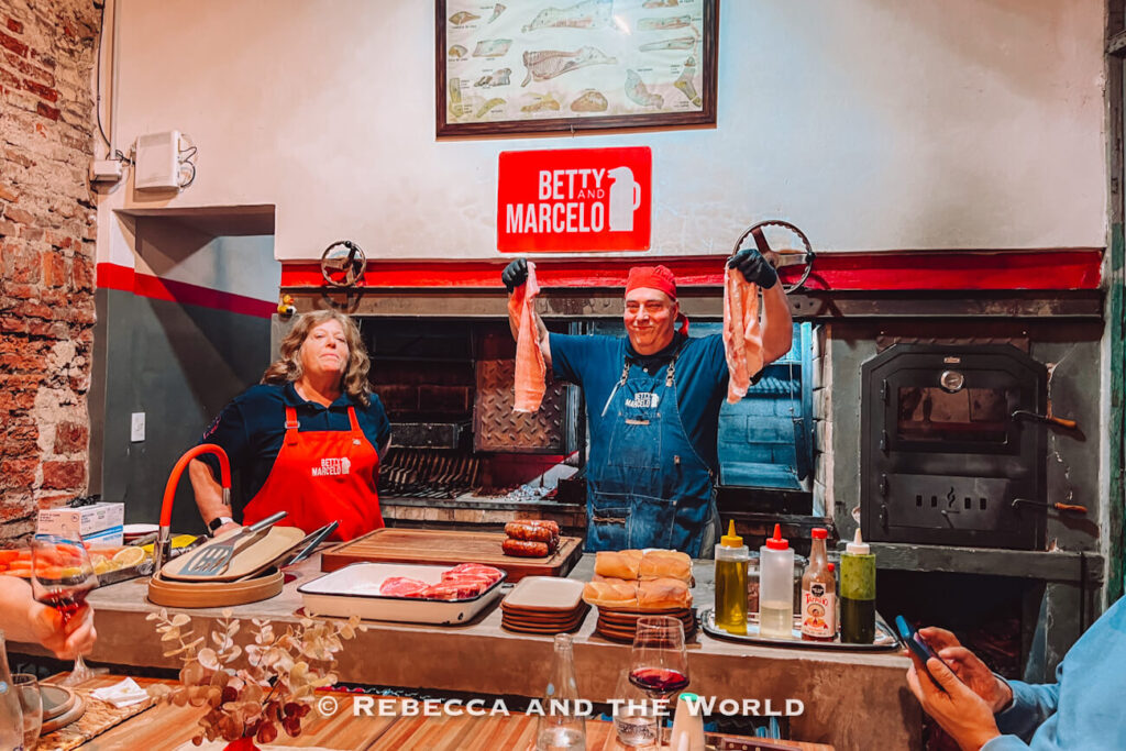 Two hosts at Betty and Marcelo Buenos Aires stand behind the grill, one holding up cuts of raw meat, presenting ingredients for a family asado in Buenos Aires.