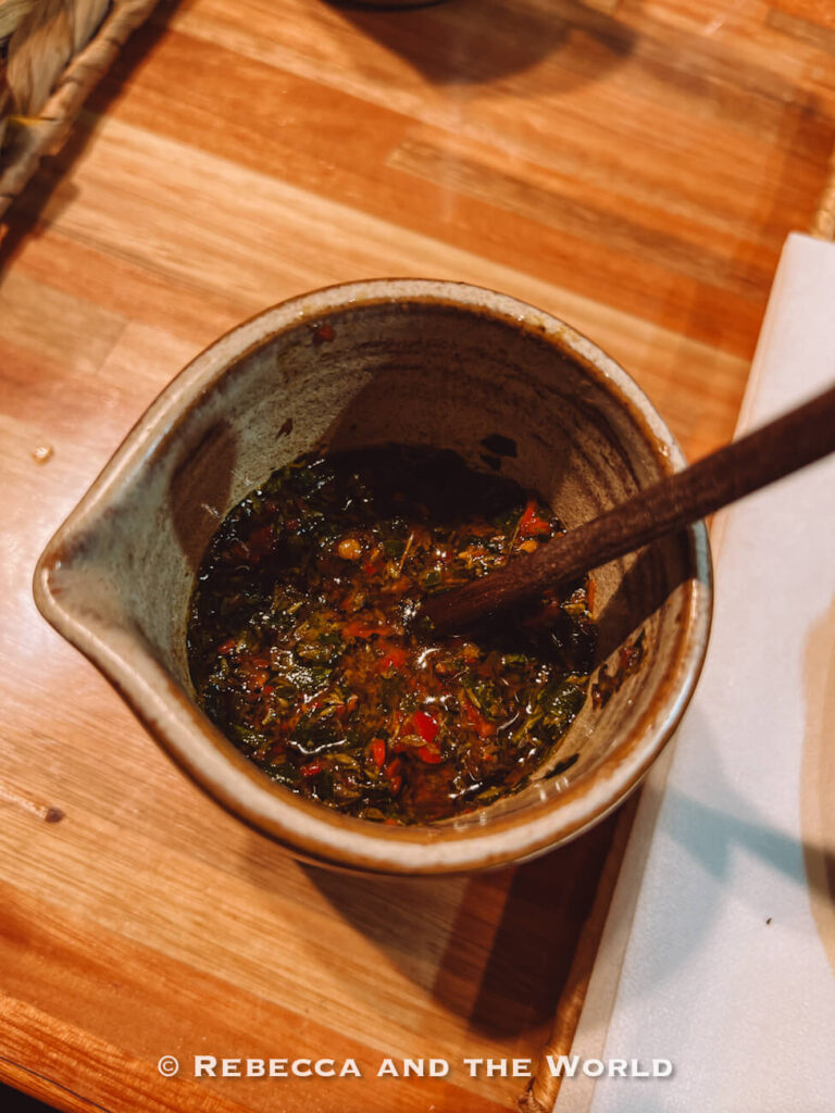 A bowl of chimichurri sauce with herbs, oil, and spices, placed on a wooden table as part of the meal at Betty and Marcelo Buenos Aires.