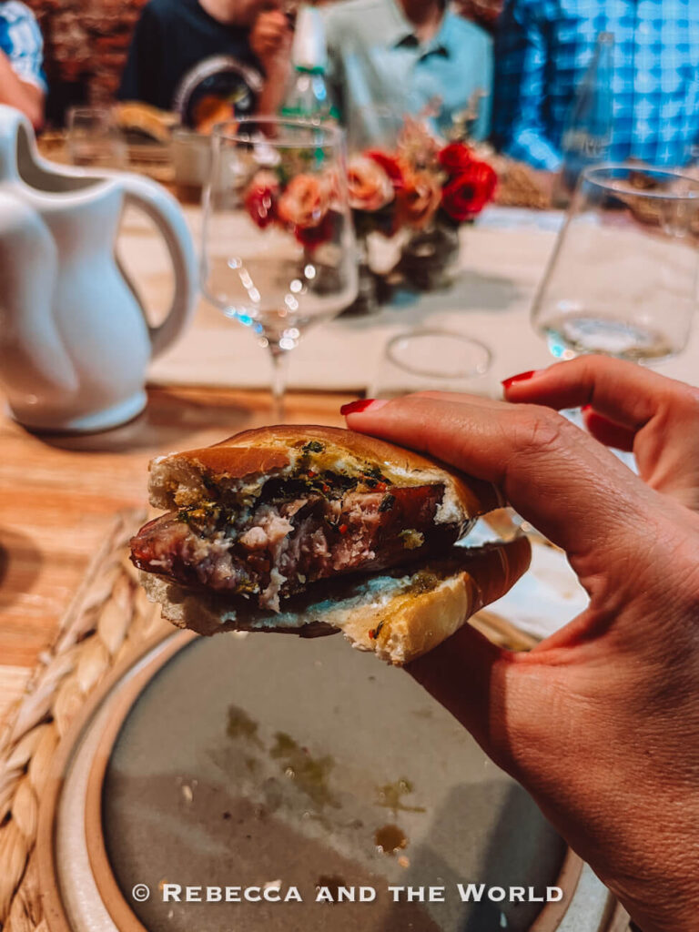 A hand holding a partially eaten choripan filled with grilled chorizo and chimichurri sauce, showing a close-up of the food enjoyed during a family asado in Buenos Aires at Betty and Marcelo.