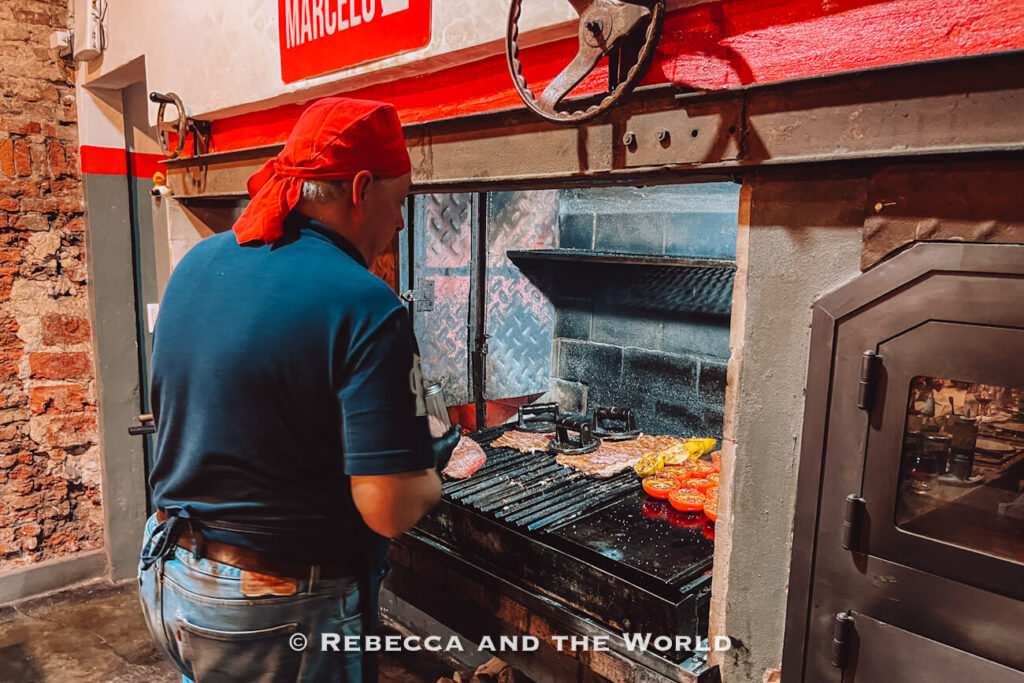 A man tending the grill at Betty and Marcelo Buenos Aires, flipping meat over open flames in a rustic kitchen setting, illustrating a traditional family asado in Buenos Aires.