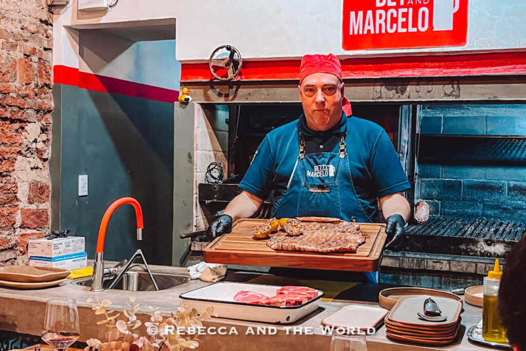 A chef at Betty and Marcelo Buenos Aires holds a wooden board with freshly grilled steak and roasted vegetables, presenting the dish in an open kitchen during a traditional Buenos Aires asado experience.