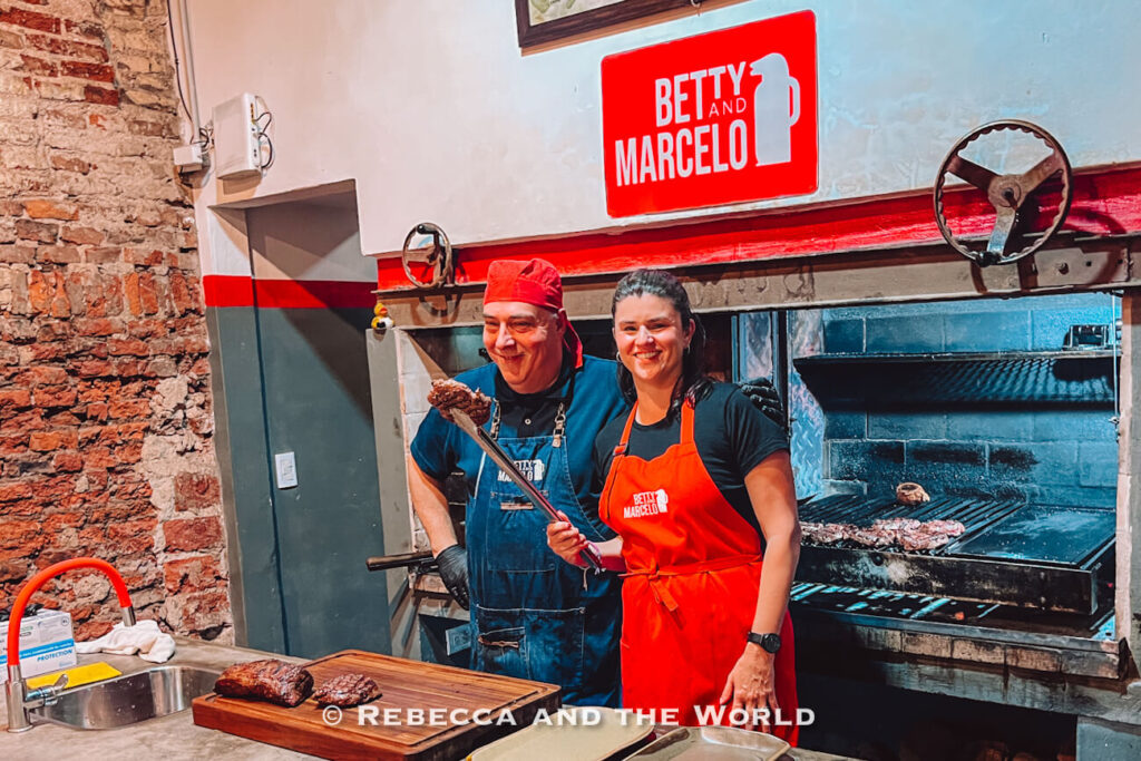 The author of this article poses with Marcelo at Betty and Marcelo Buenos Aires, with grilled meat resting on a cutting board and the open grill behind them during a family asado in Buenos Aires.