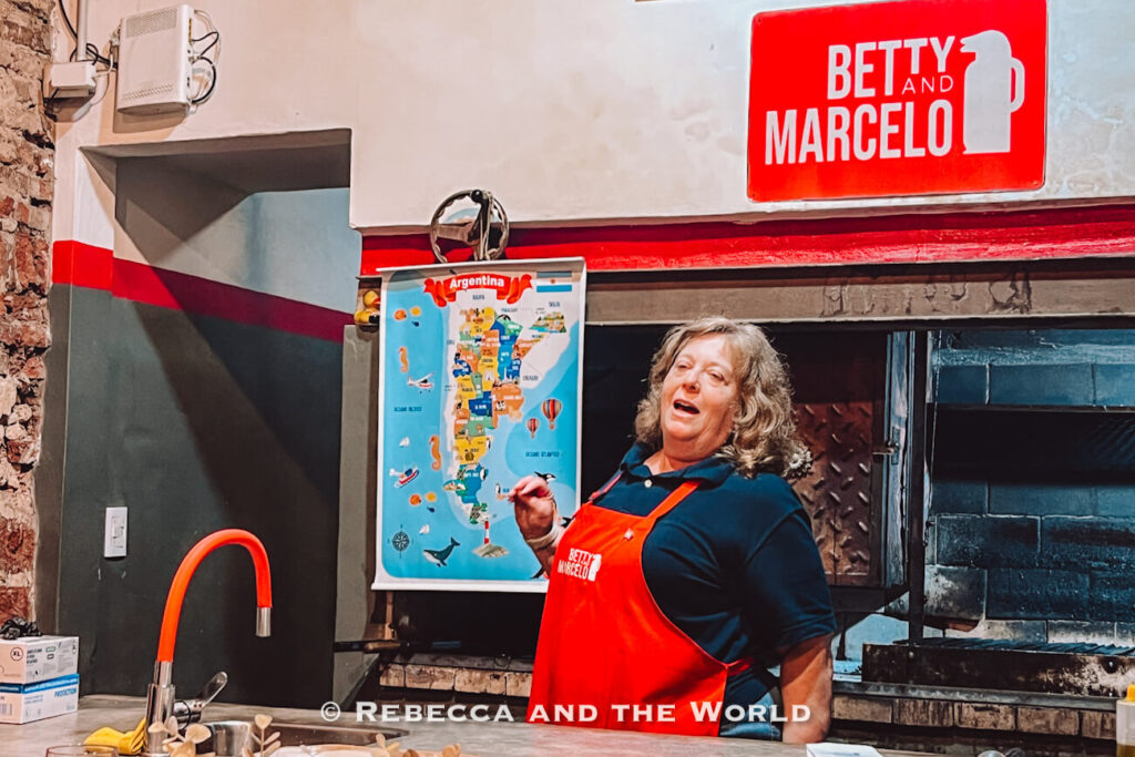 A woman wearing a red apron at Betty and Marcelo Buenos Aires speaks to guests near the grill, with a map of Argentina behind her, adding to the interactive feel of a family asado in Buenos Aires.