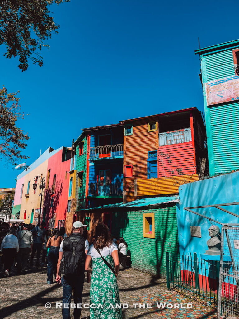 Visitors walk along a cobblestone street lined with brightly painted buildings in vivid blues, reds, and yellows. This iconic Buenos Aires neighborhood offers free sightseeing and photo opportunities for budget travelers.