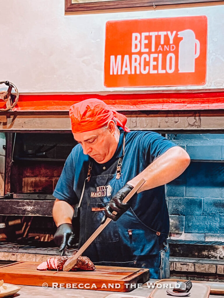 A chef wearing a red headscarf and apron slices grilled meat on a wooden board inside a restaurant kitchen, with a sign reading “Betty and Marcelo” behind him. This scene reflects local parrilla culture, a key experience when planning a Buenos Aires travel budget.