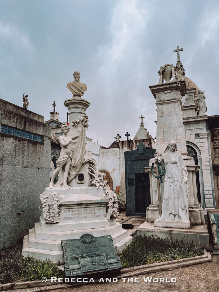 Ornate marble statues and mausoleums stand closely together in a historic cemetery under a cloudy sky. The Recoleta Cemetery is an interesting place to visit in Buenos Aires - but you will need to factor the cost into your daily expenses.