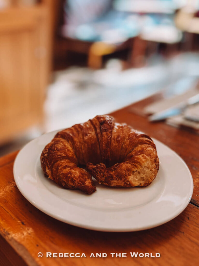 A golden, flaky medialuna sits on a white plate on a wooden café table, with a softly blurred background. A simple, affordable breakfast option often found in Buenos Aires cafés, ideal for budget-friendly travel days.