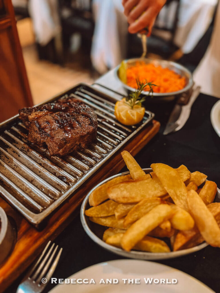 Juicy grilled steak served on a hot metal grill with a side of thick-cut fries and a small dish of mashed vegetables, with a hand reaching in the background. A classic Buenos Aires dining experience, highlighting how food can be a major part of a Buenos Aires travel budget.