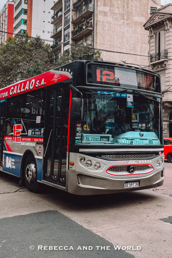 A city bus with route number 12 displayed on the front drives through an urban street lined with apartment buildings. Local buses are one of the cheapest transportation options in Buenos Aires, ideal for keeping travel costs low.