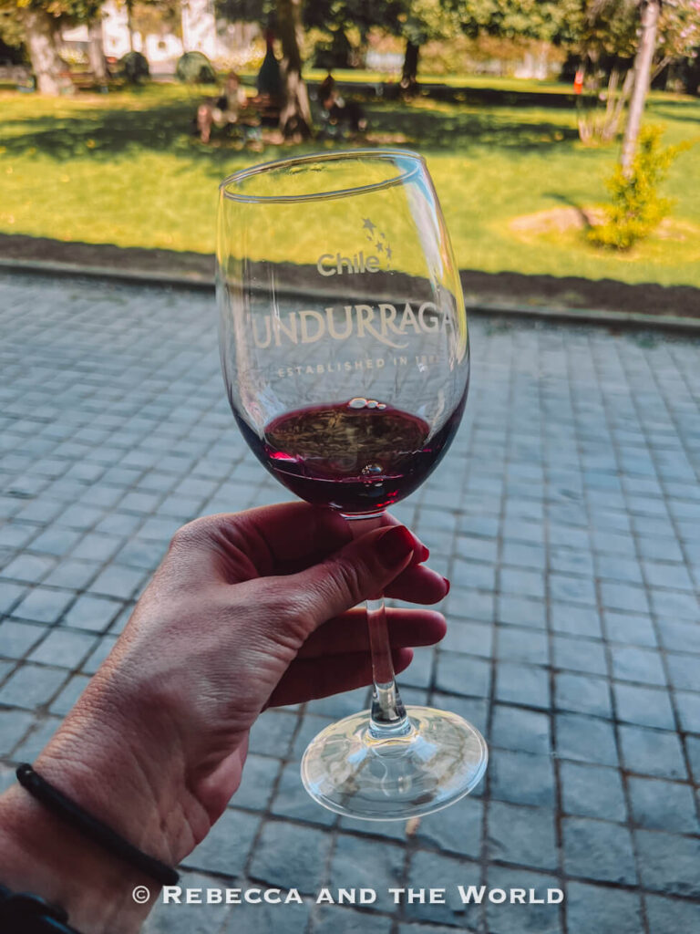 A hand holds a glass of red wine labeled “Undurraga,” with a garden blurred in the background, illustrating a relaxed wine tasting experience in the Maipo Valley.