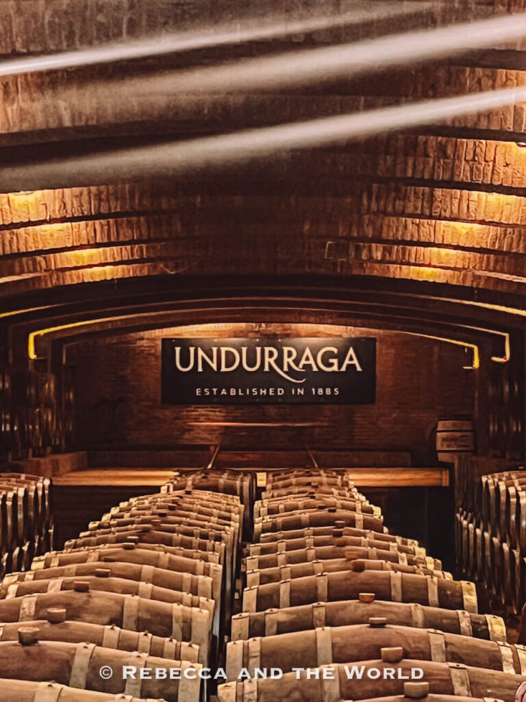 Rows of wooden barrels line a dimly lit cellar with a sign reading “Undurraga,” showcasing part of a Maipo Valley tour focused on wine production and aging.
