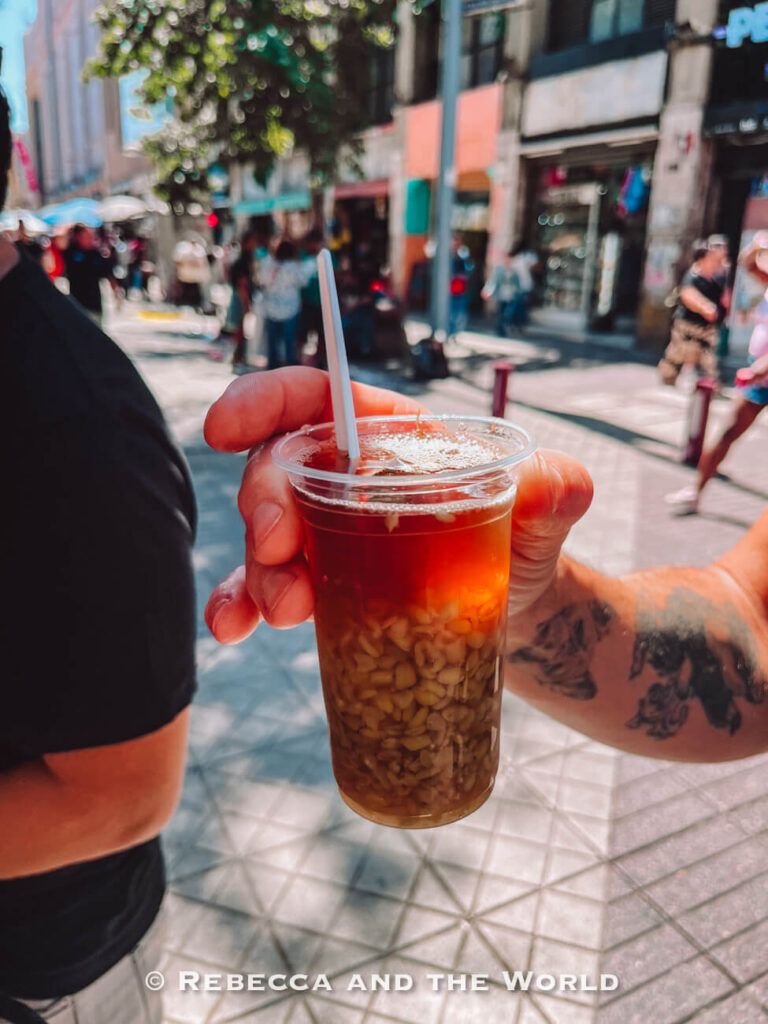 A plastic cup of mote con huesillo held on a city street, a classic Chilean drink often included in a Santiago food tour highlighting traditional Chilean food.
