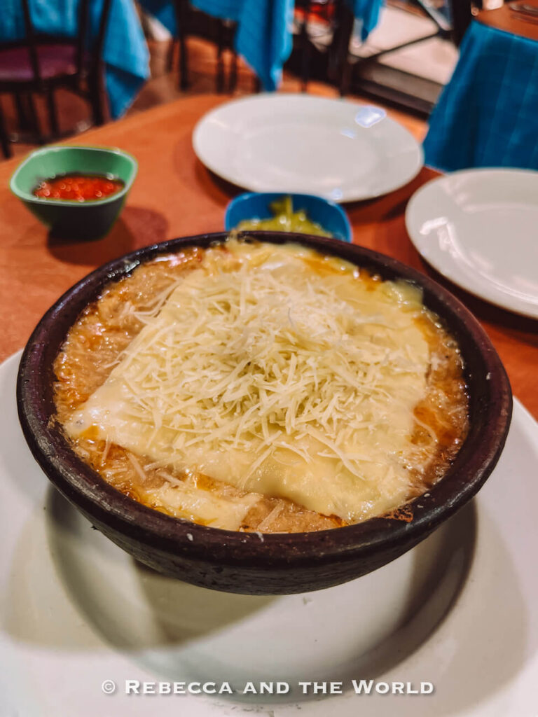 A clay bowl of cheesy baked Chilean dish - chupe de jaiba - served at a restaurant table, representing comforting traditional Chilean food you might try on a Santiago market food tour.