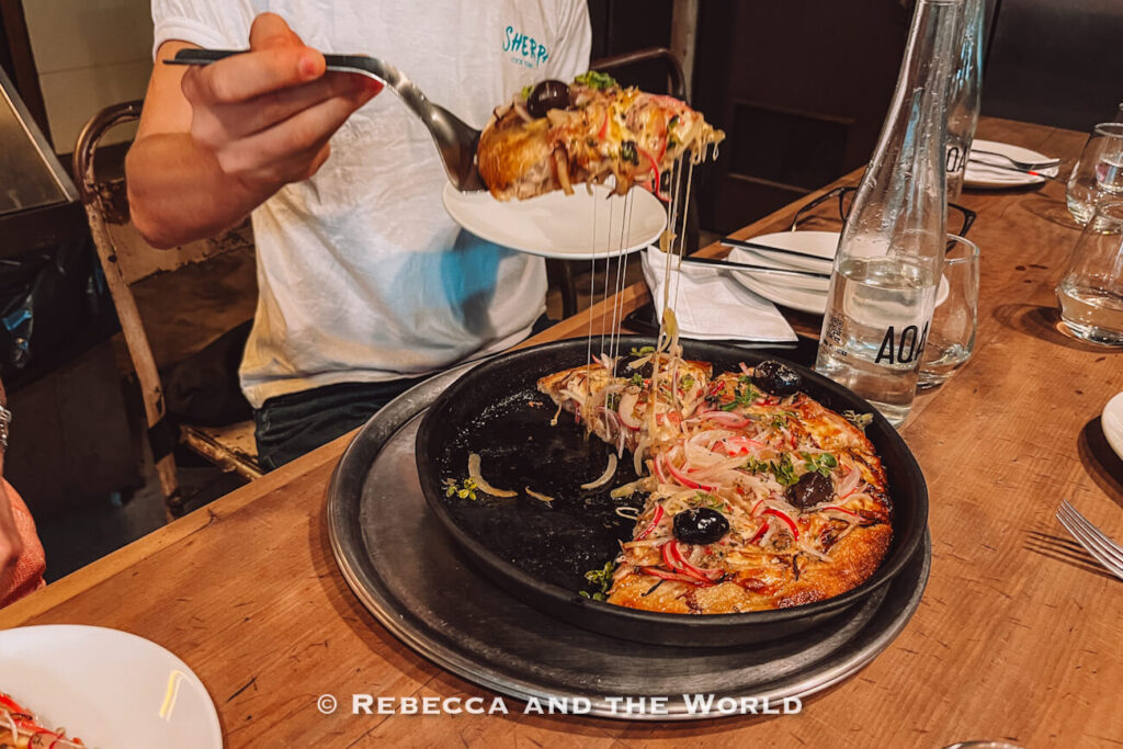 A person lifting a slice of pizza with melted cheese stretching from the pan, topped with onions and olives, during a Sherpa food tour Buenos Aires at a casual restaurant table.